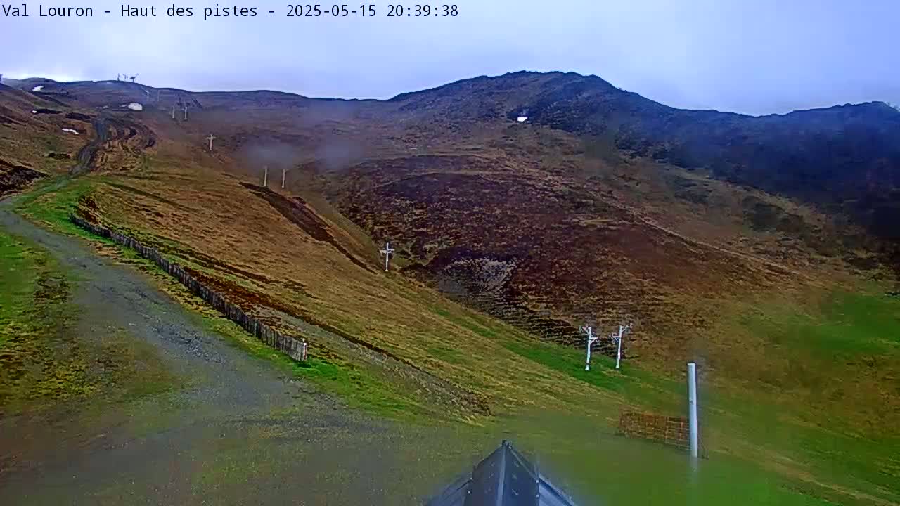 A mostly cloudy day reveals a mountainside with patches of green grass and brown earth, a dirt road, and a ski lift.
