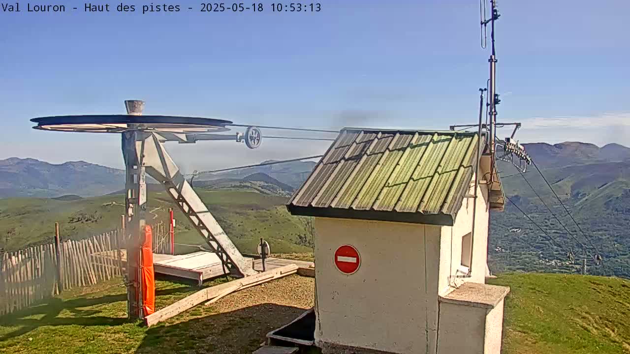 A ski lift's pulley system and control building are situated atop a grassy hill overlooking a mountainous valley under a clear, sunny sky.