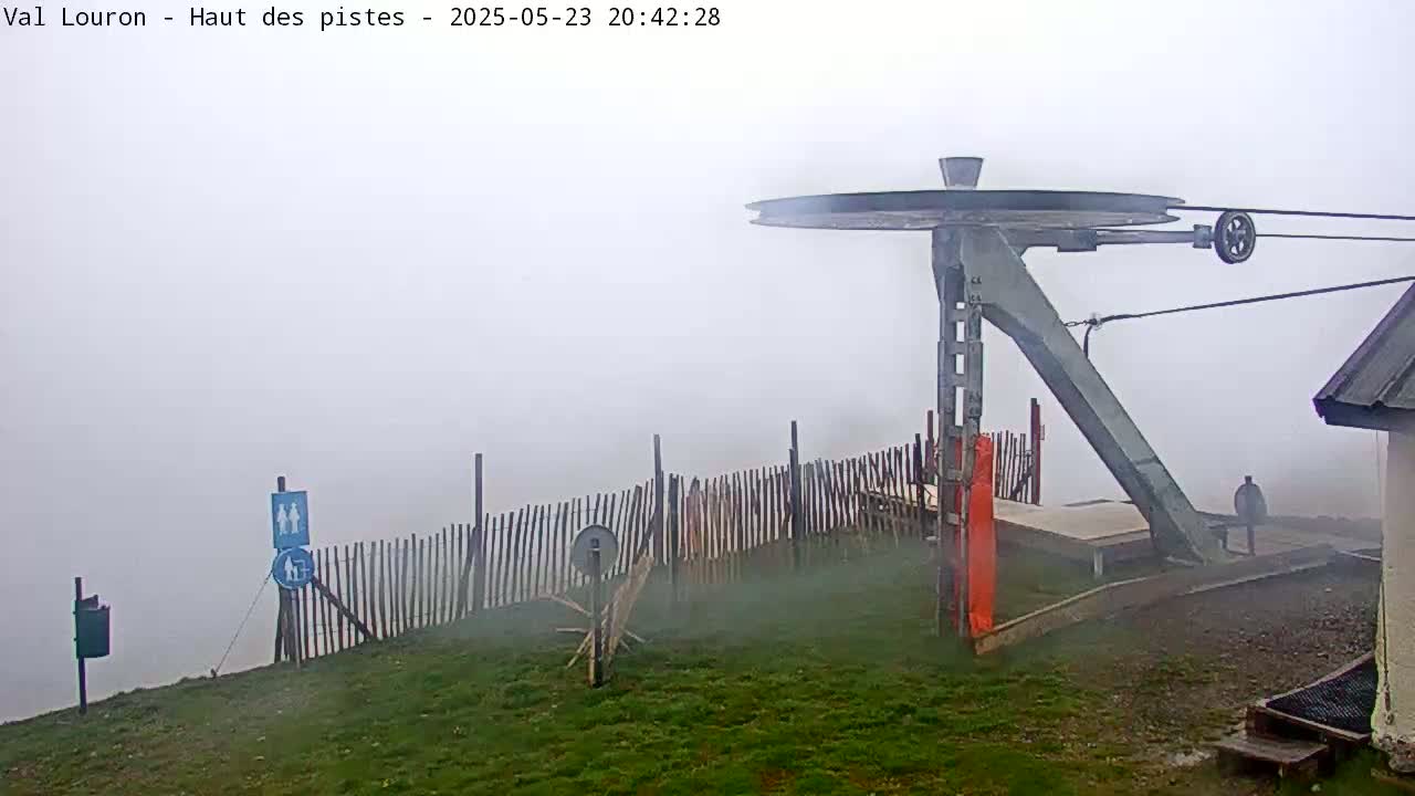 A ski lift pulley system is visible through a thick fog, with a wooden fence and grassy area surrounding it.