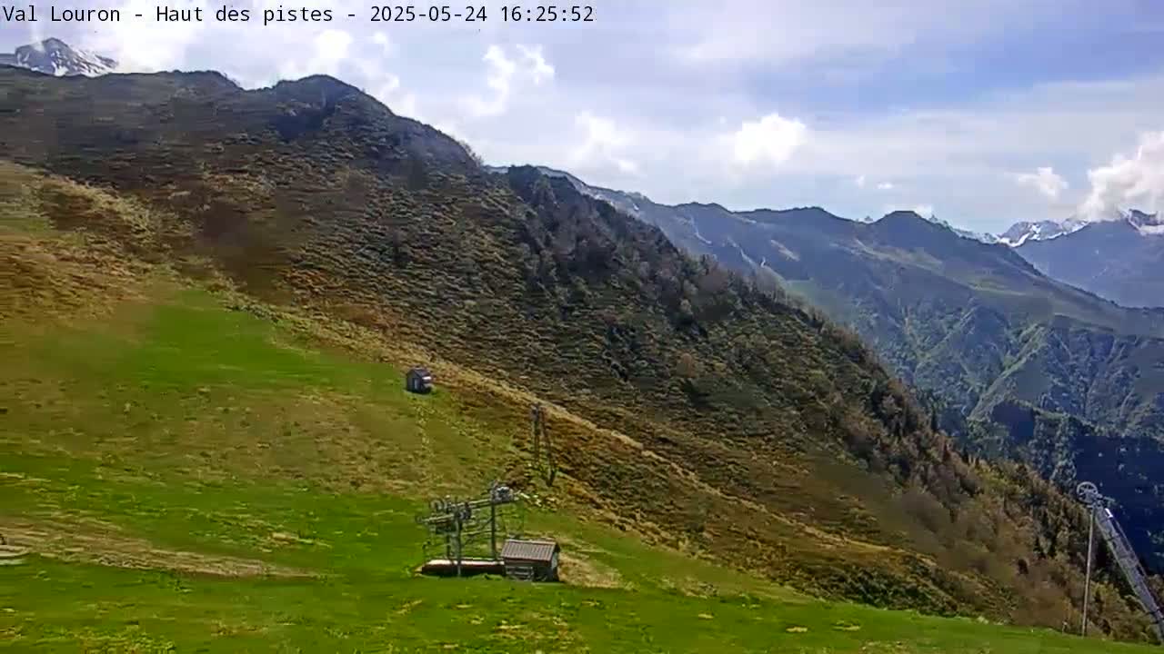 A mostly sunny day reveals a grassy mountain slope with a ski lift and small buildings, overlooking a vast, mountainous valley.