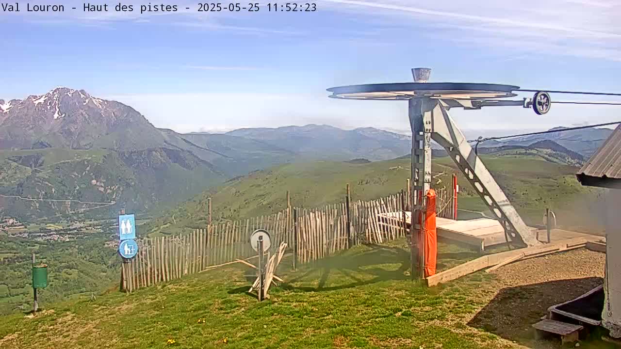A mountaintop view on a partly sunny day shows a ski lift pulley system, a wooden fence, and distant snow-capped mountains.
