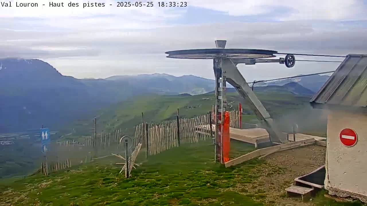 A mountaintop ski lift pulley system and building are visible under a cloudy sky, overlooking a grassy valley and distant mountains.