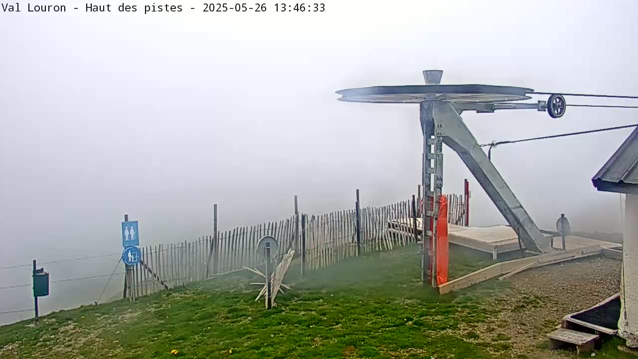A ski lift pulley system is visible in foggy conditions, surrounded by a grassy area, a wooden fence, and a small building.