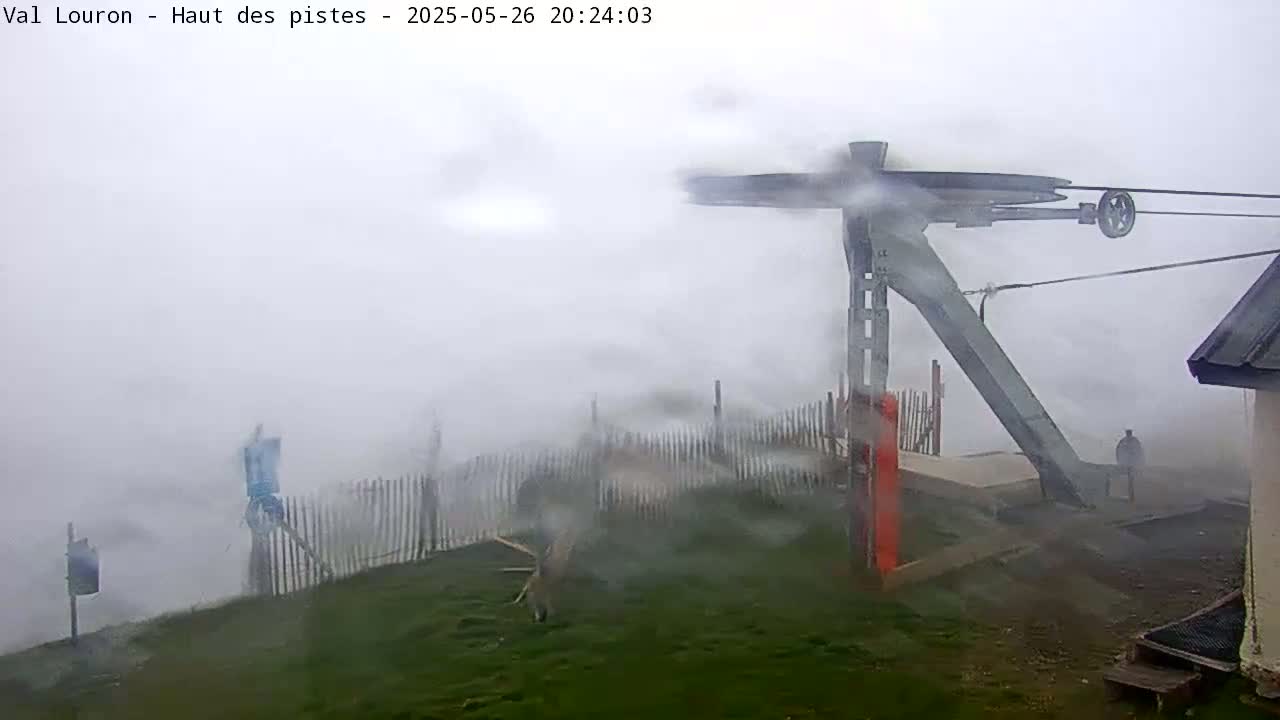 A ski lift's pulley system is partially obscured by heavy fog, with a grassy area and a wooden fence visible in the foreground.