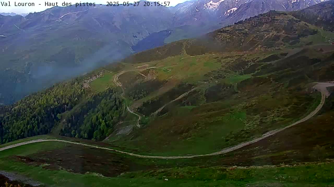 A winding dirt road snakes through a mountainous landscape under a hazy sky, with snow-capped peaks in the distance.
