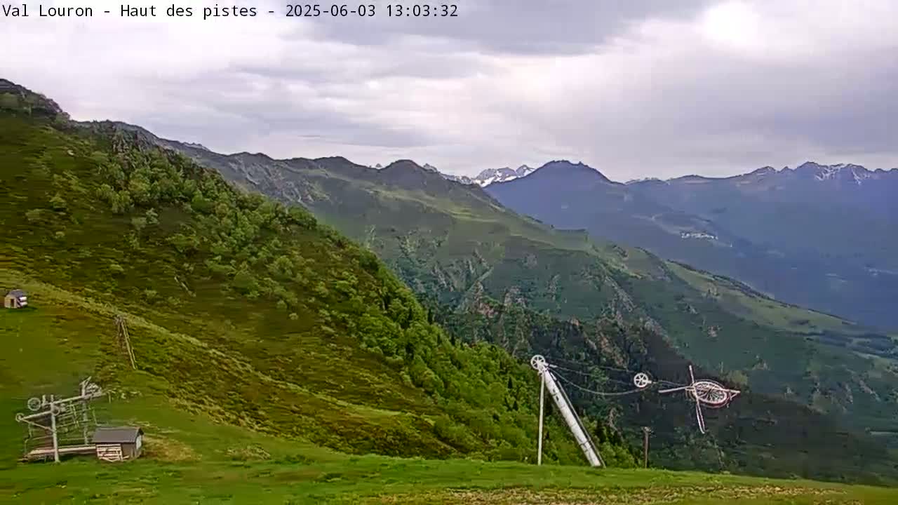 A mostly cloudy day reveals a green mountainside with some small buildings and ski lift equipment, overlooking a valley and distant, snow-capped mountains.