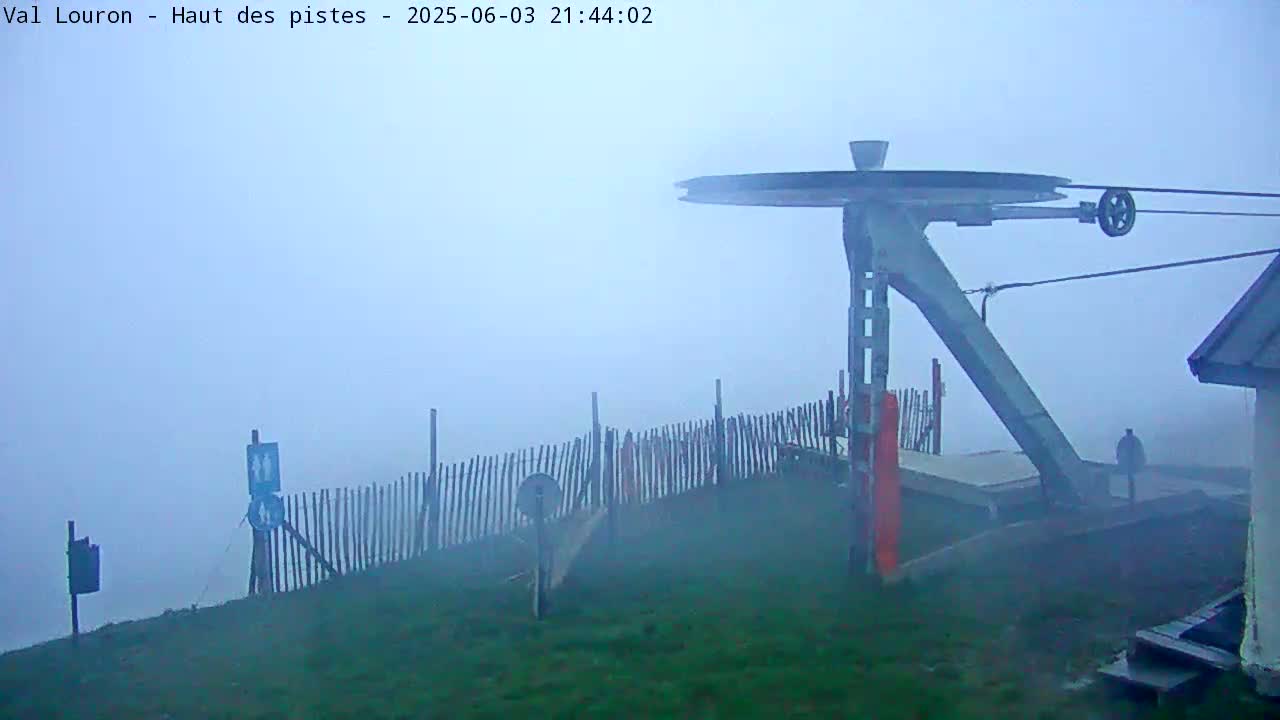 A ski lift mechanism is visible in foggy conditions, partially obscured by mist, with a wooden fence and grassy area surrounding it.
