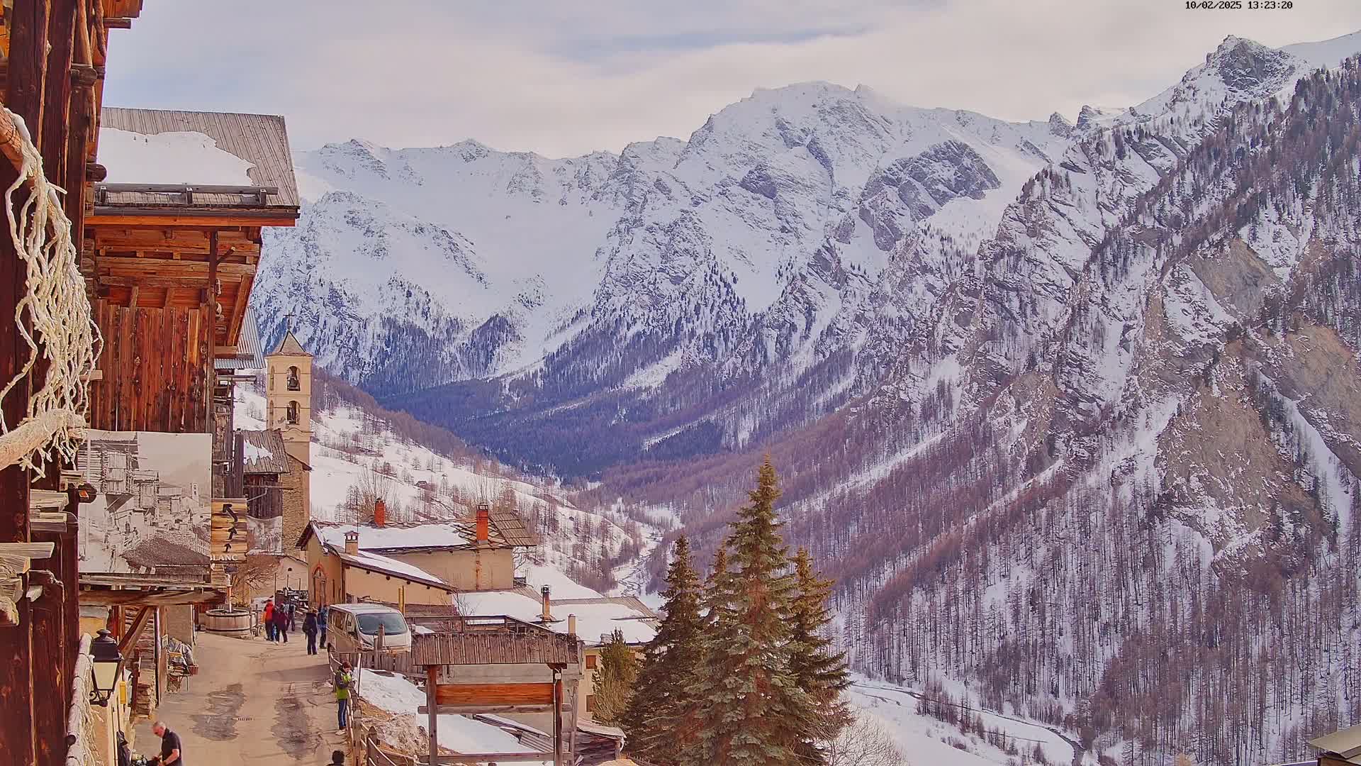 A snow-covered mountain village with a church and several buildings is nestled in a valley between snow-capped mountains under a cloudy sky.