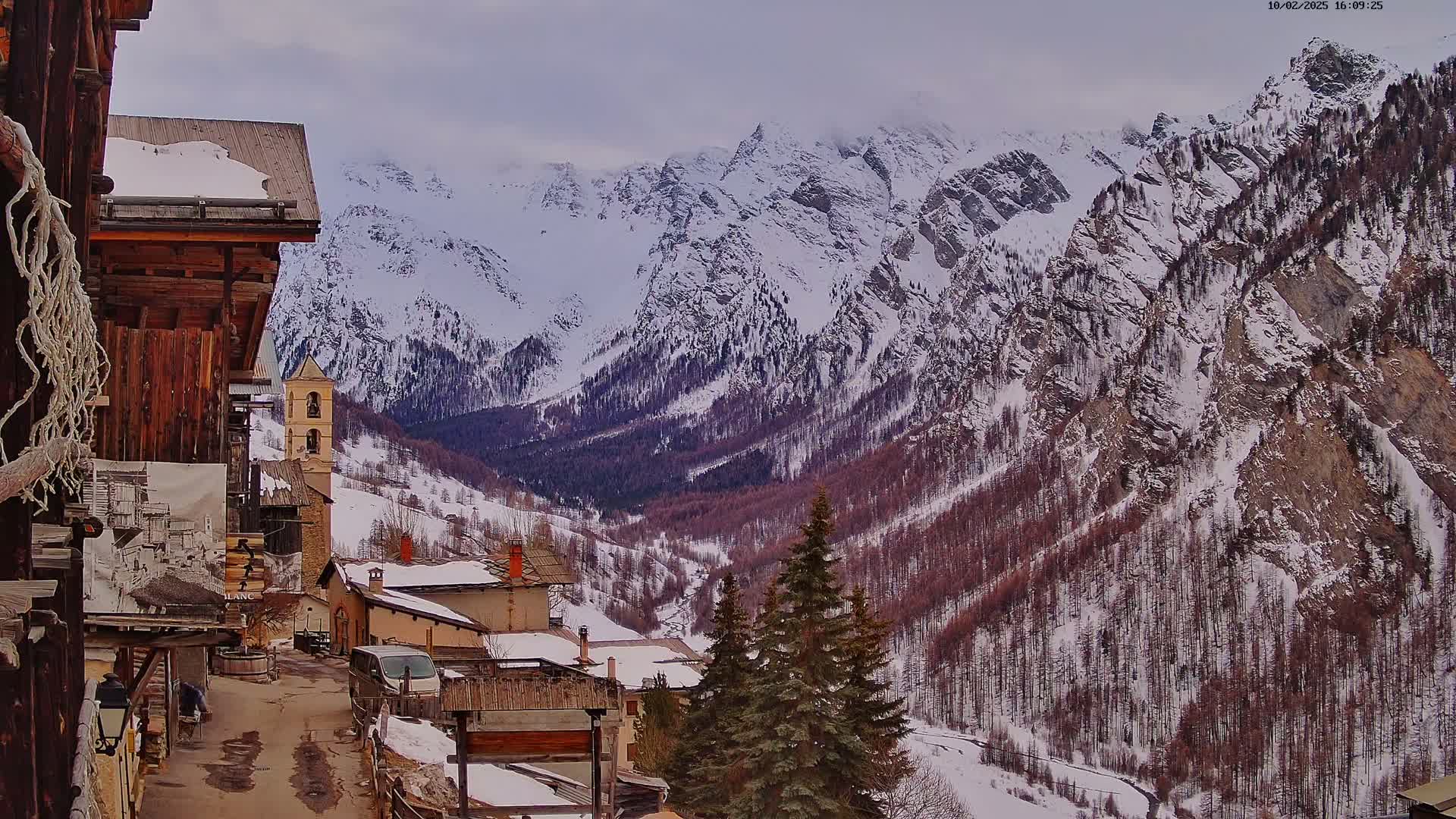 A snow-covered mountain valley village is viewed from a wooden building's perspective under an overcast sky.