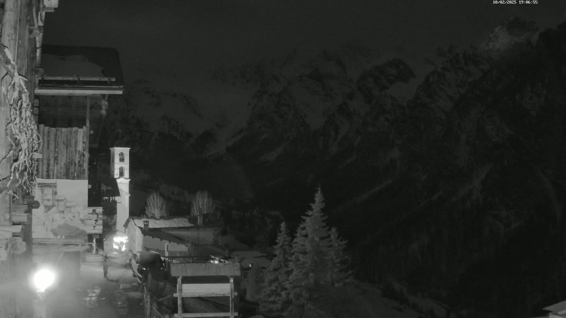 A nighttime, snow-covered mountain range is visible behind a small village with a bell tower, viewed from a building's exterior.
