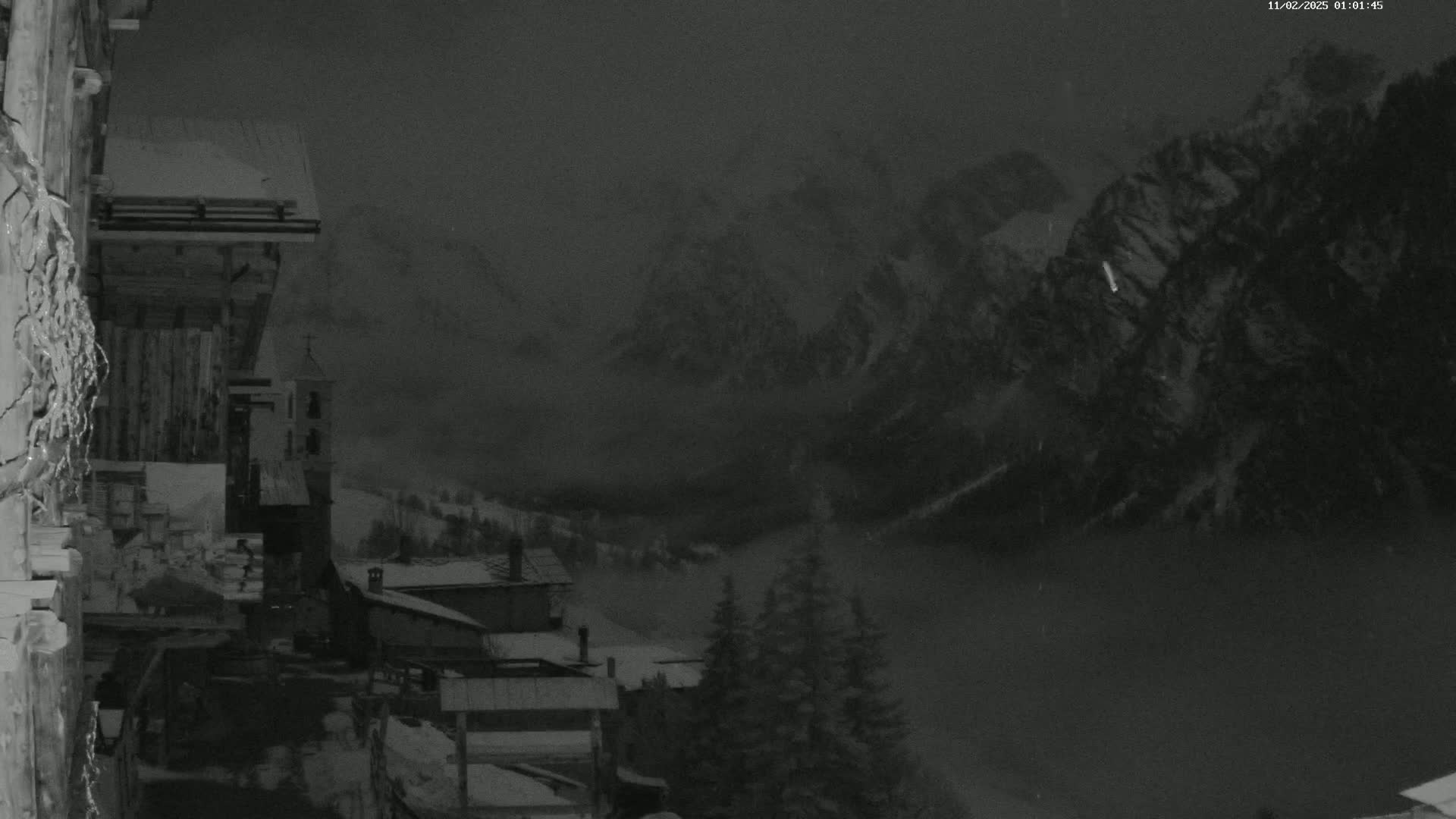 A snow-covered village nestled in a valley between dark, snow-capped mountains is shrouded in fog under a nighttime sky.