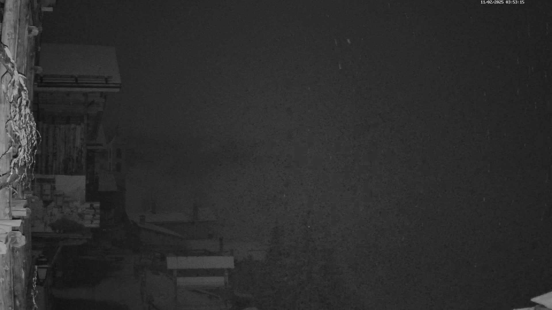 A nighttime, snowy scene shows a snow-covered building and surrounding structures under a dark sky.