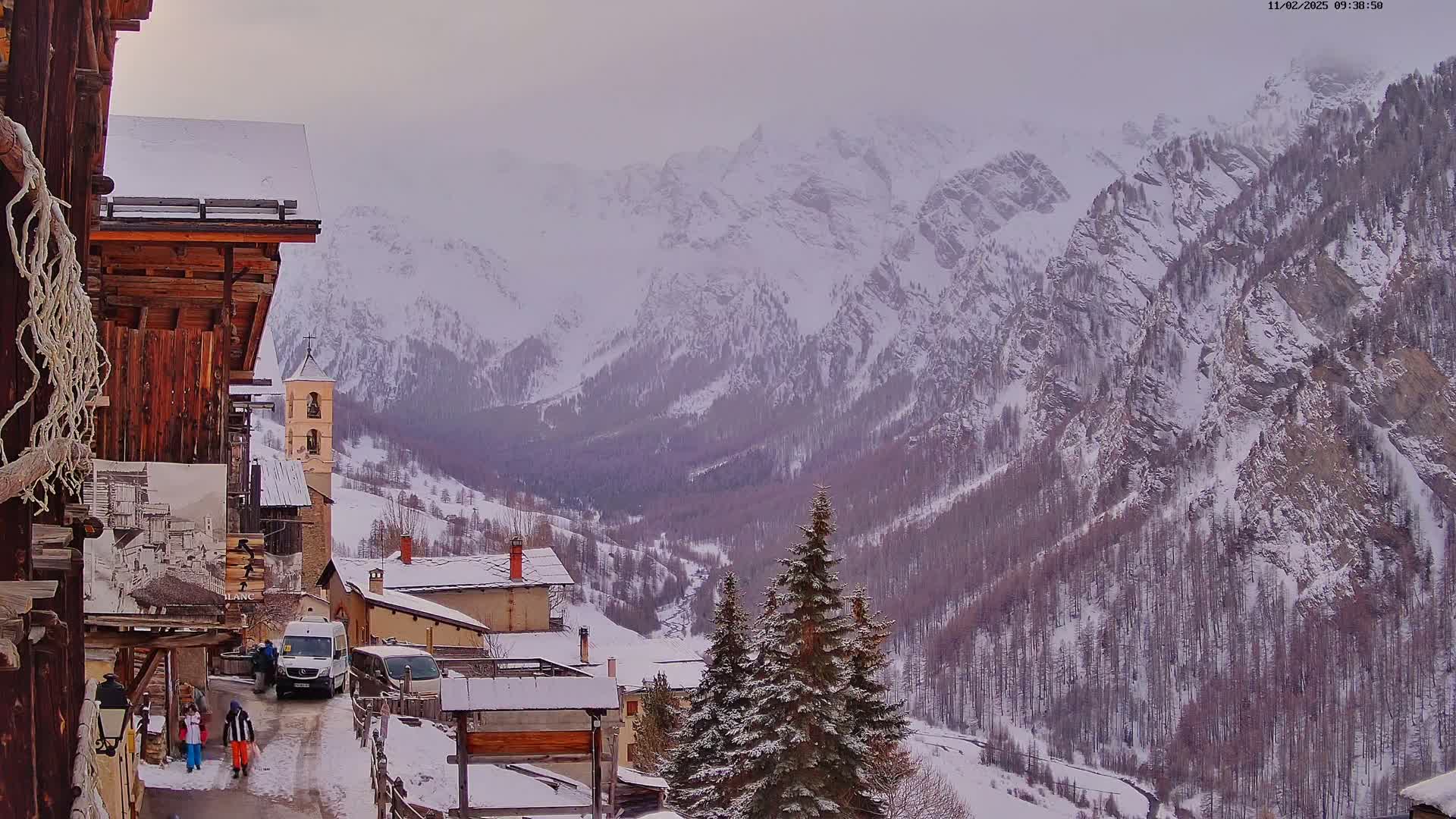 A snow-covered village nestled in a valley between towering, snow-capped mountains on an overcast day.