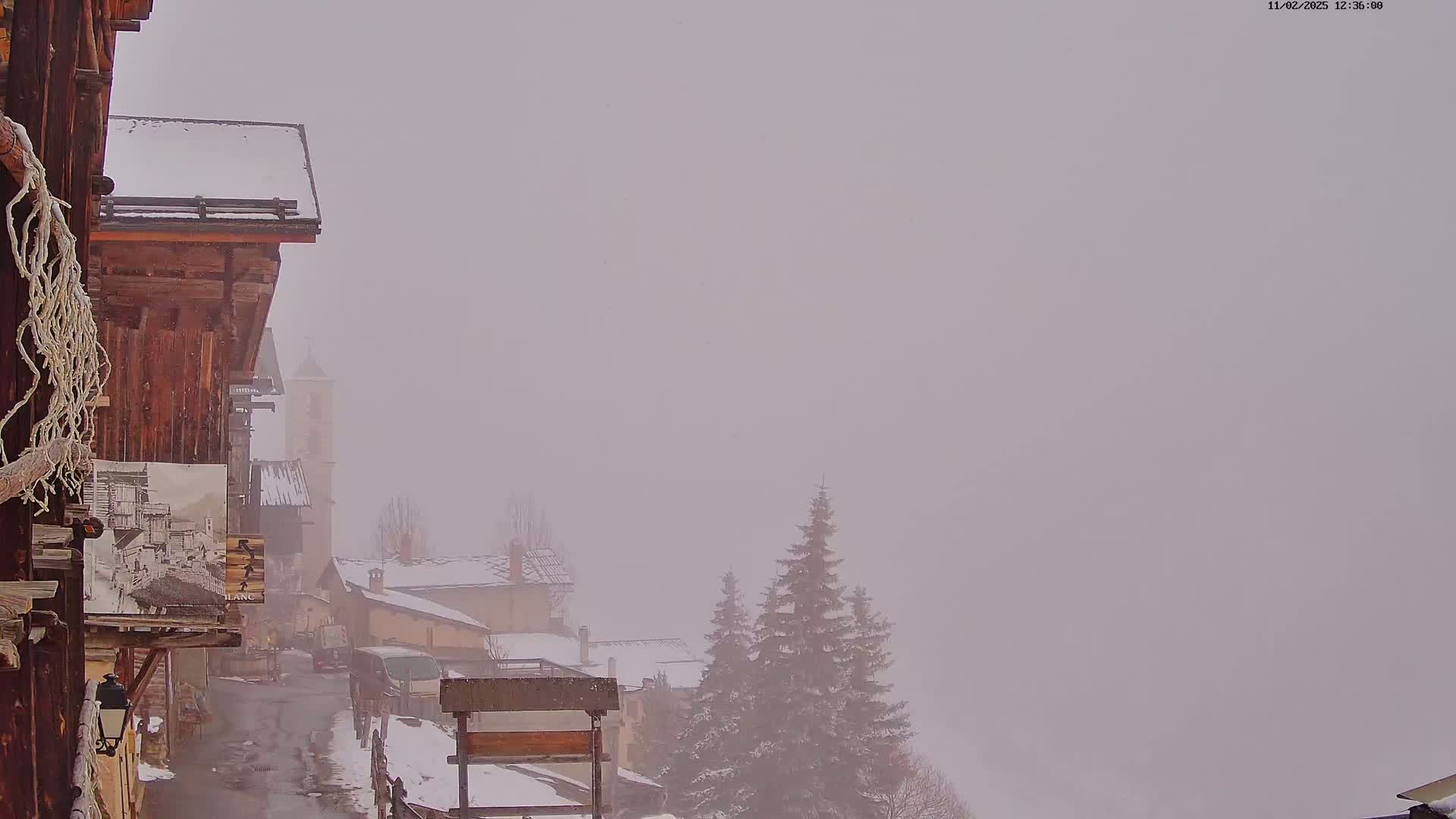 A snow-covered village is shrouded in heavy fog, with snow-dusted evergreens visible in the distance.