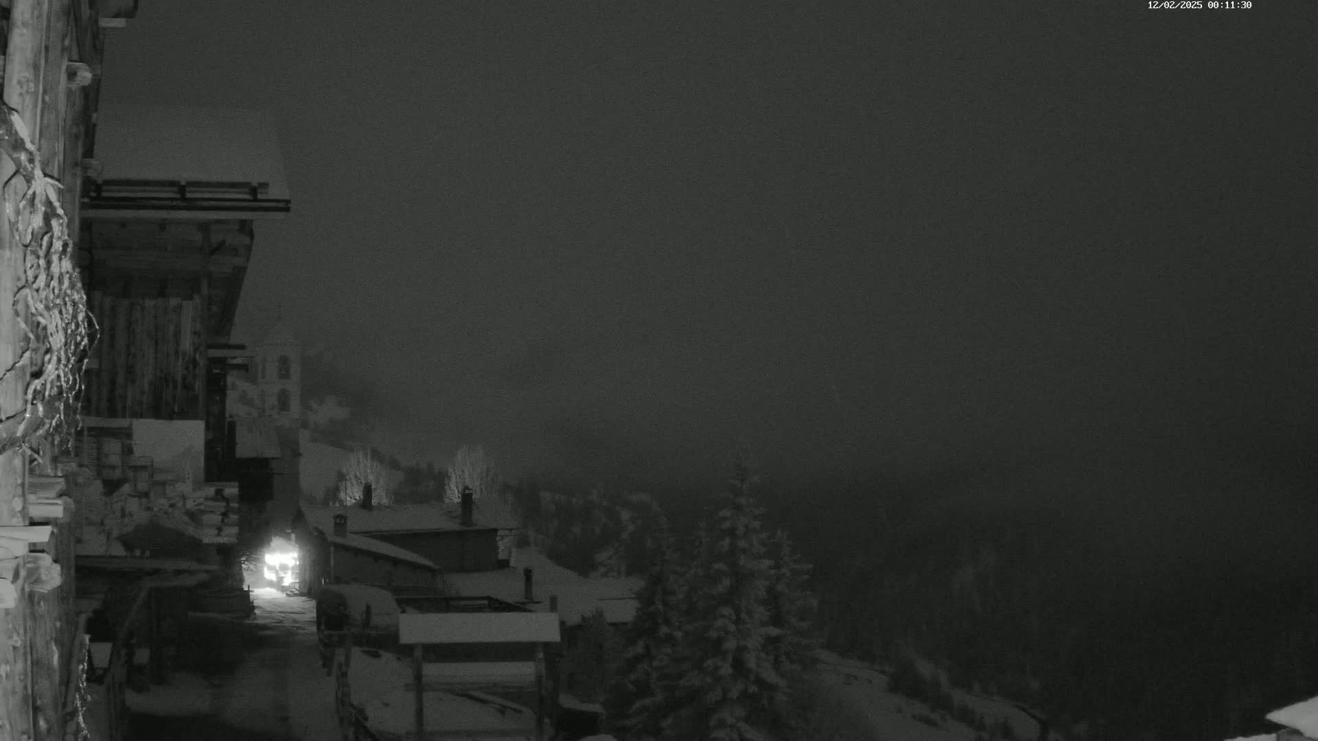 A nighttime, snowy scene shows a snow-covered village nestled in a foggy, mountainous region, with a vehicle's headlights visible on a road below.