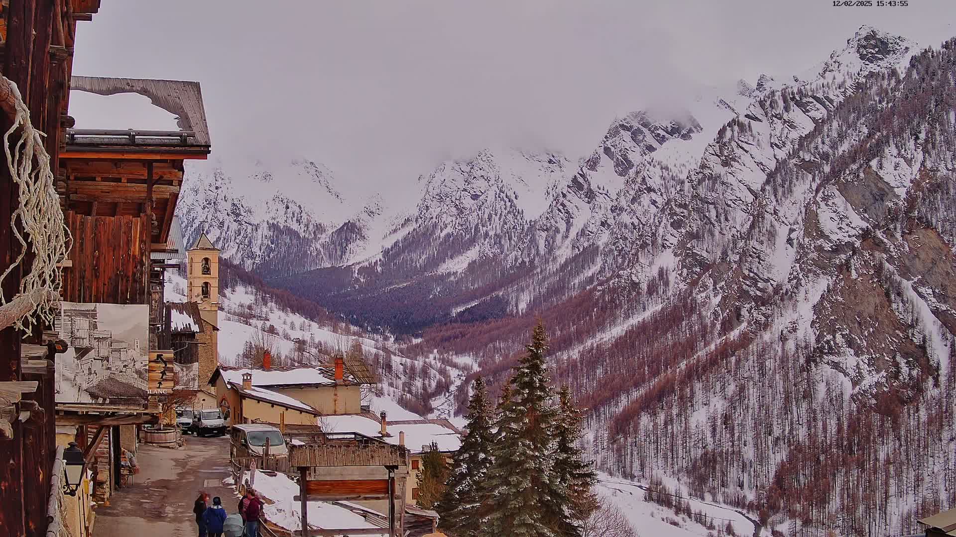 A snow-covered mountain village nestled in a valley between towering, snow-capped mountains under an overcast sky.