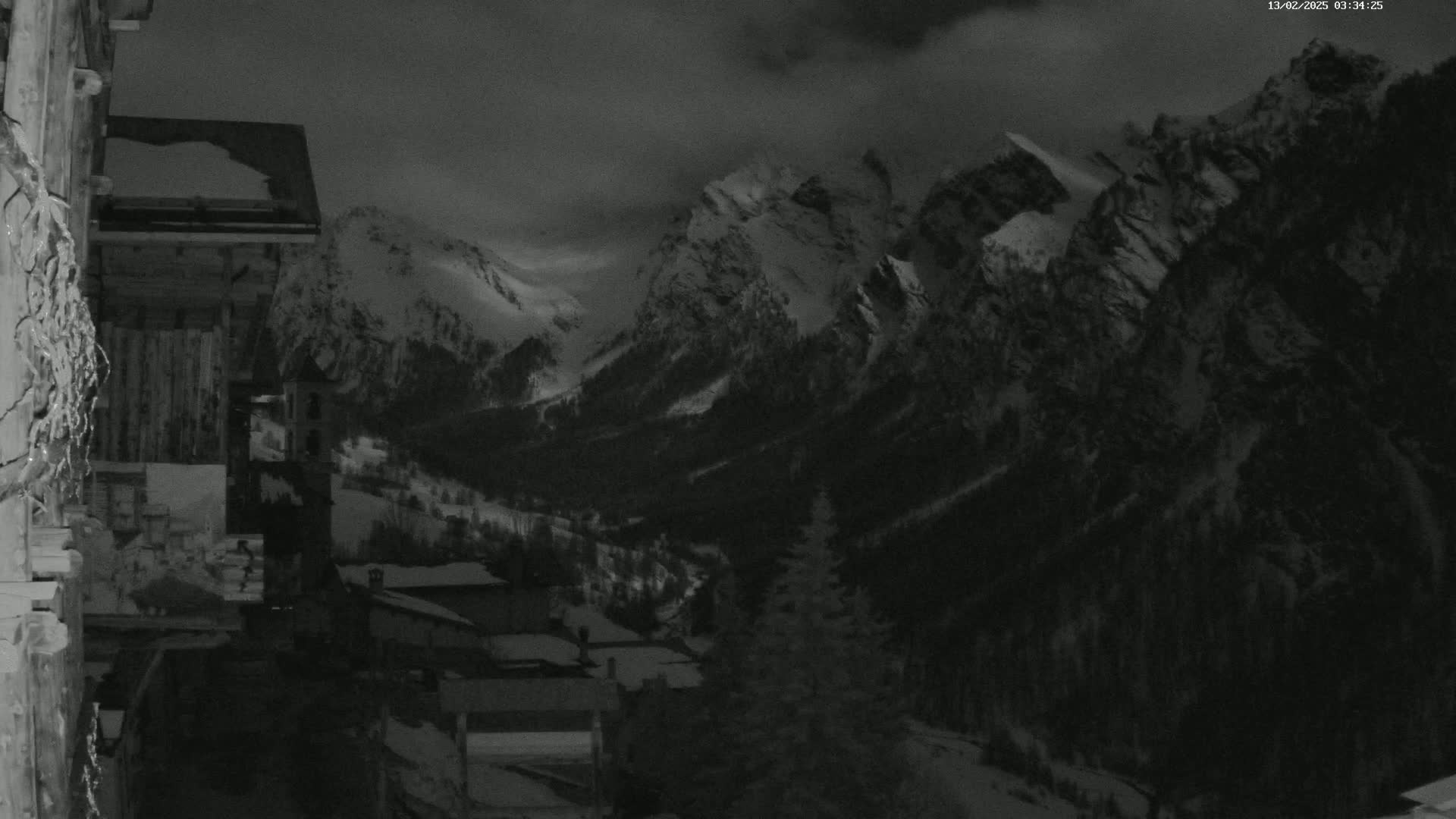 A nighttime, snow-covered mountain range is visible from a village nestled in the valley below, under a cloudy sky.