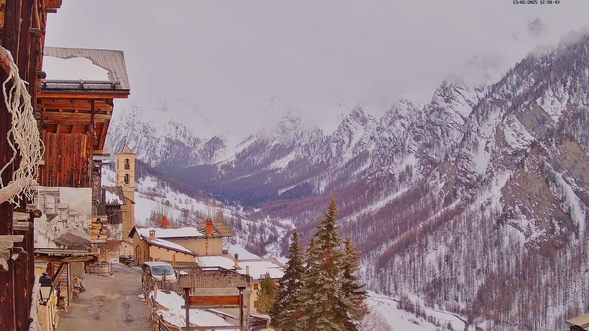 A snow-covered mountain village with a church bell tower is nestled in a valley surrounded by snow-capped mountains under an overcast sky.
