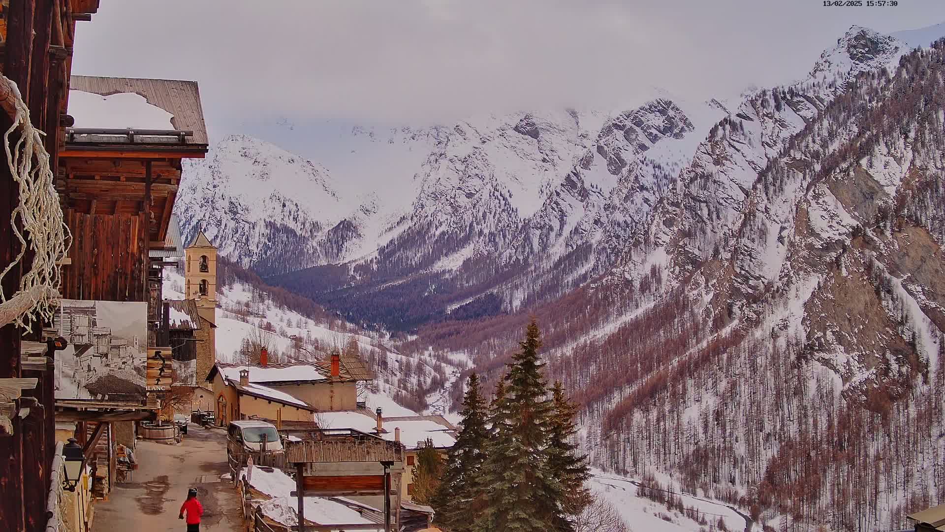 A snow-covered village nestled in a valley, overlooked by towering, snow-capped mountains under a cloudy sky.