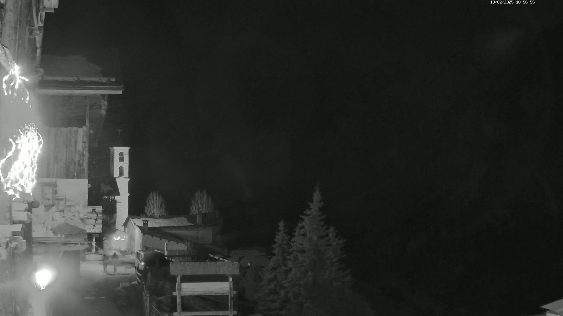 A nighttime, snow-covered village scene features a church bell tower, some buildings, and evergreens under a dark sky.