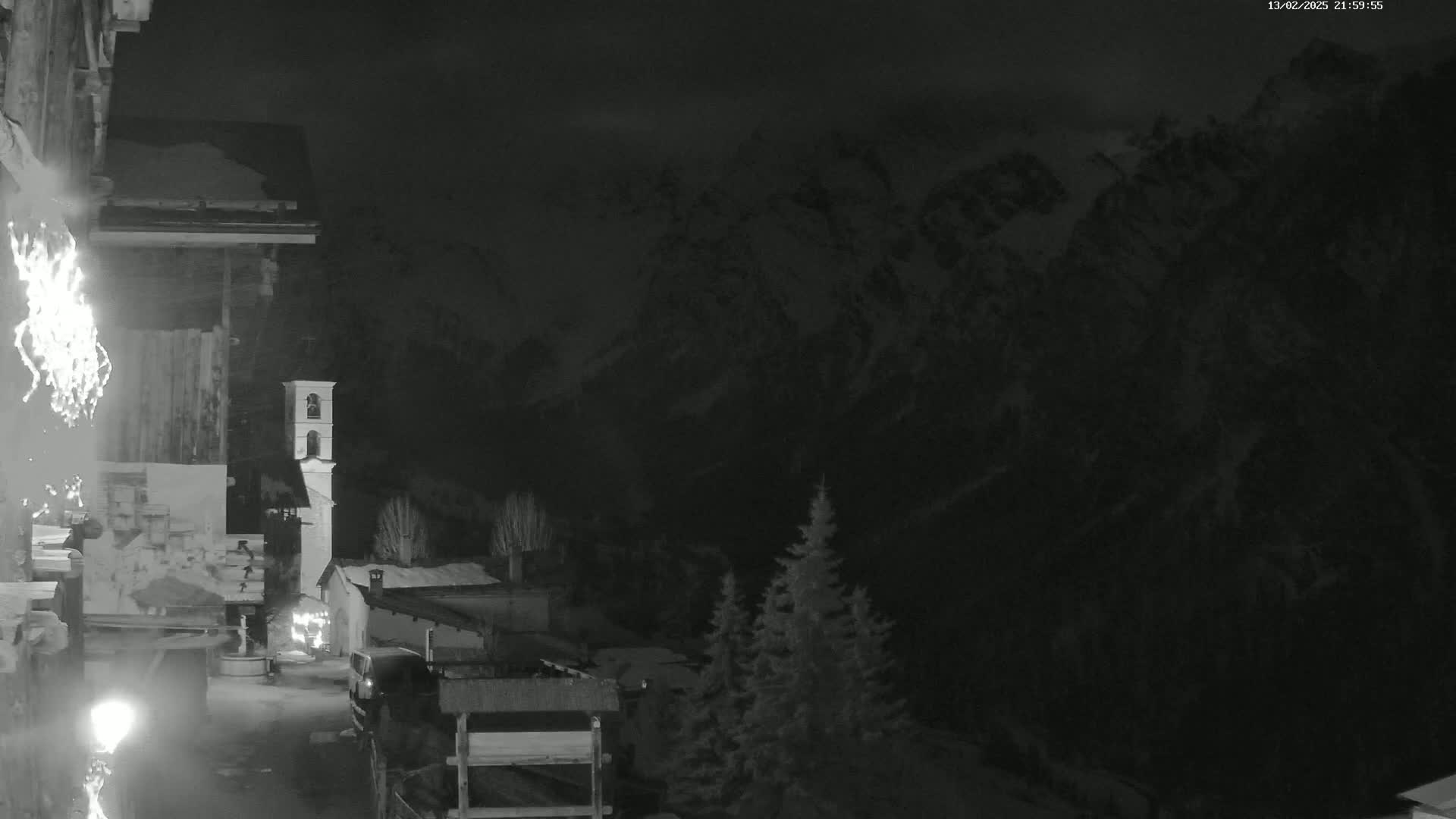 A nighttime, snow-covered mountain range is visible from a village containing a bell tower and several buildings, under a dark, cloudy sky.