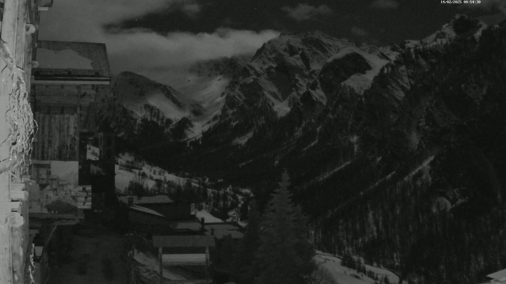 A nighttime, snowy mountain valley scene is viewed from a wooden building, with cloudy skies overhead.