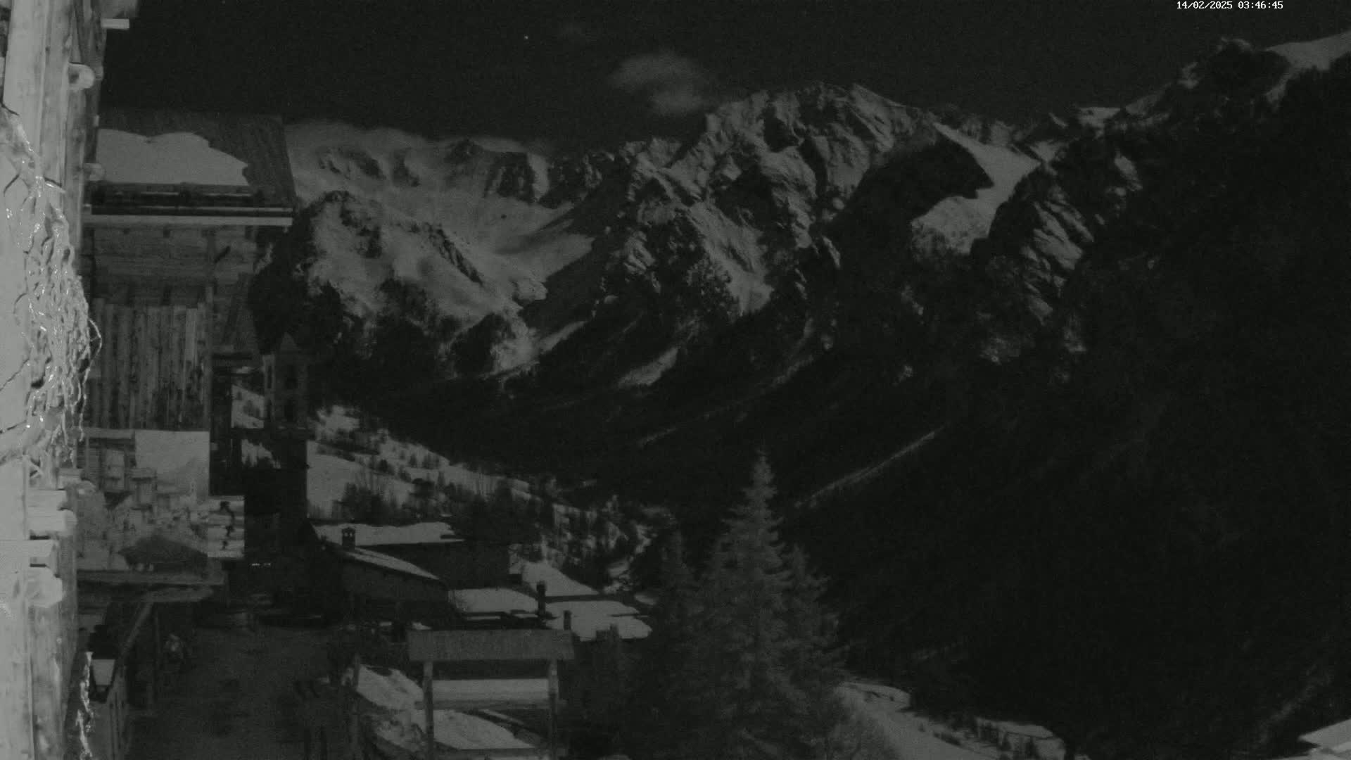 A nighttime view from a wooden building shows a snow-covered mountain range and a small village nestled in the valley below.