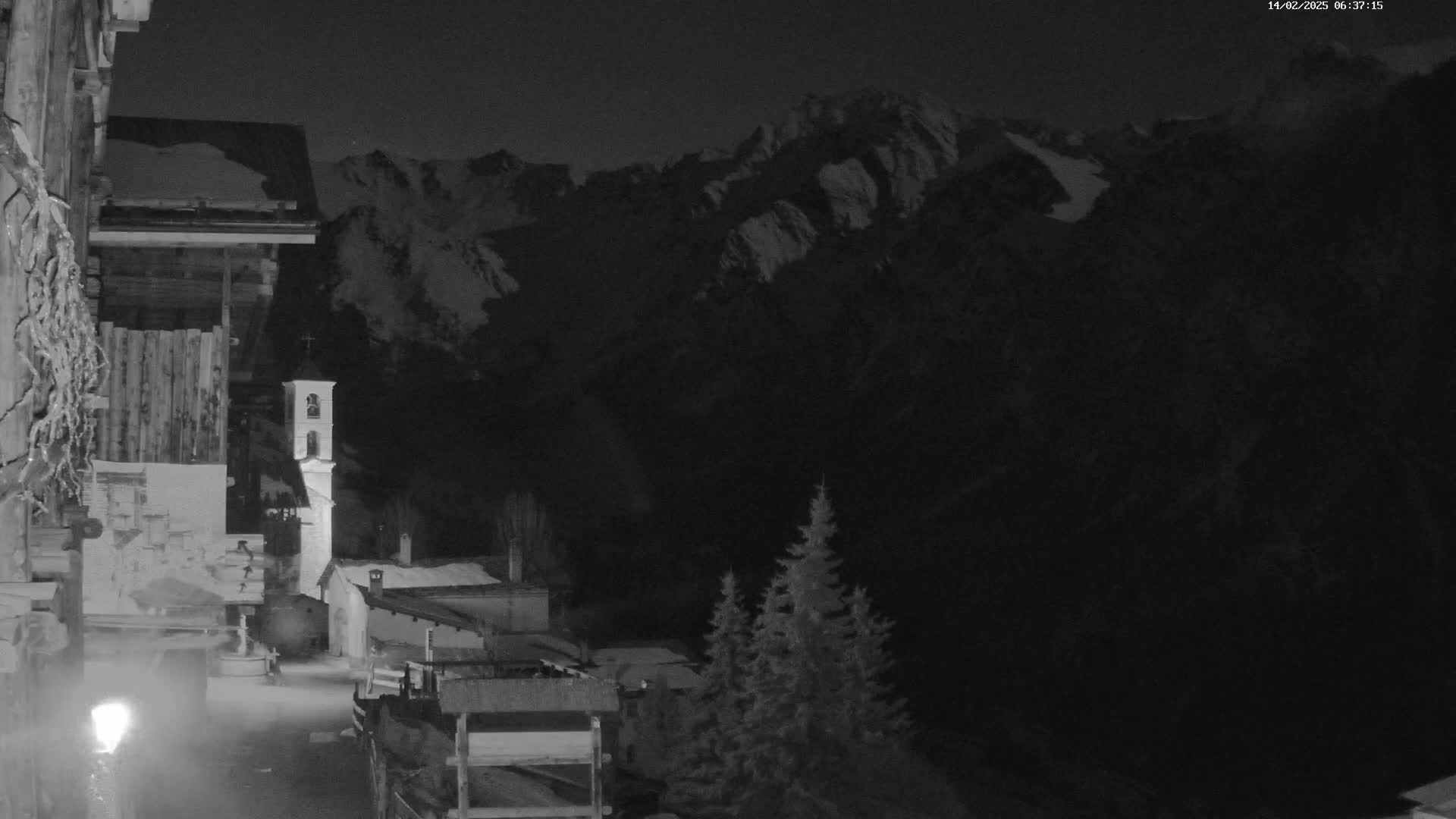 A nighttime view of a snow-dusted village nestled at the foot of a mountain range under a dark sky.
