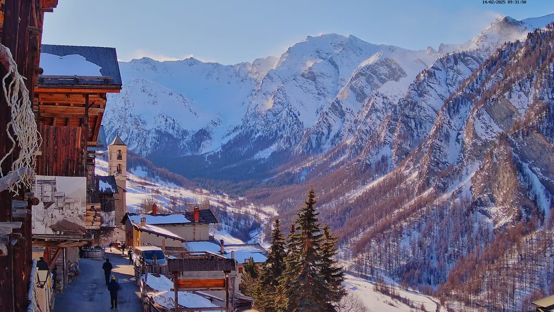 A snow-covered village nestled in a valley between tall, snow-capped mountains on a sunny day.