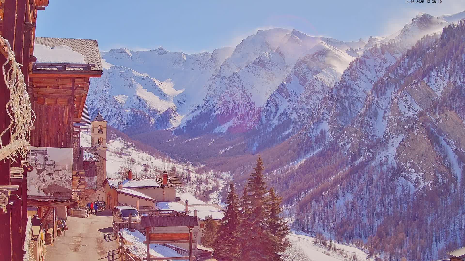 A snow-covered village nestled in a valley, overlooked by a sunlit, snow-capped mountain range under a clear blue sky.