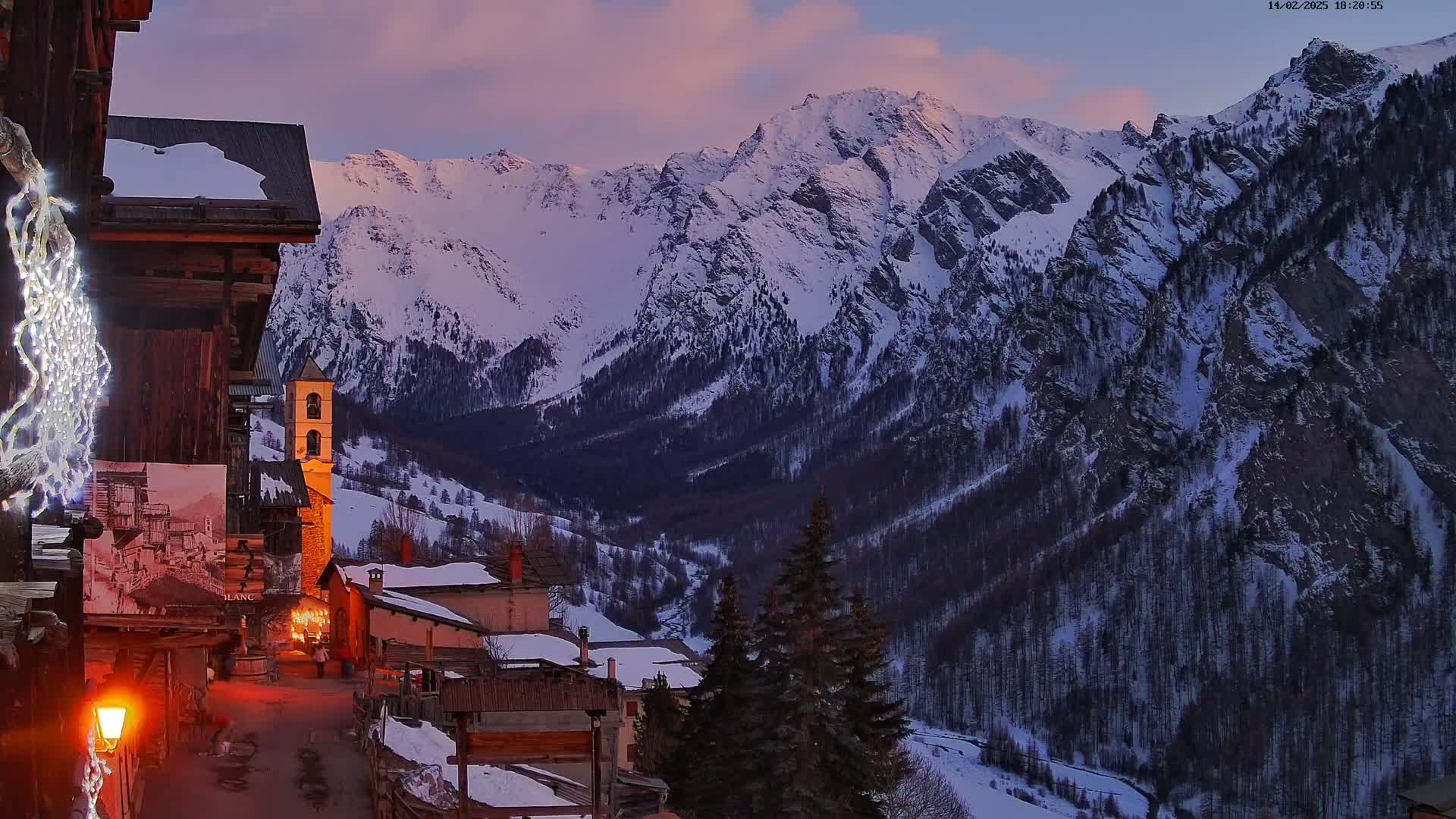 A snow-covered mountain range is seen at dusk, overlooking a small village with old stone buildings and a church steeple, illuminated by warm streetlights.