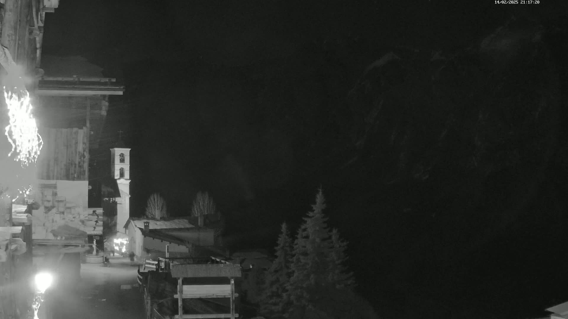 A nighttime, snow-covered village scene features a bell tower and several buildings, with dark mountains in the background under a clear night sky.