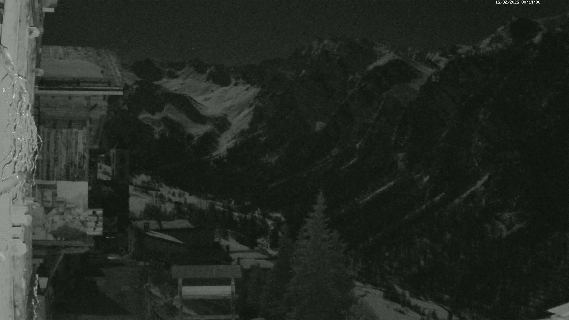 A nighttime, low-light view shows snow-covered mountains in the distance, with a few buildings and trees visible in the foreground.