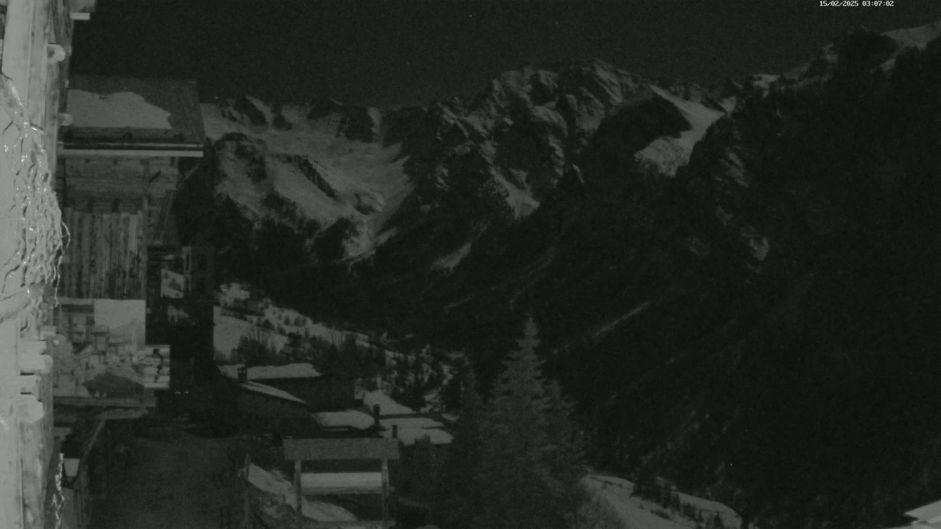 A nighttime, snow-covered mountain range is viewed from a village nestled in a valley below.