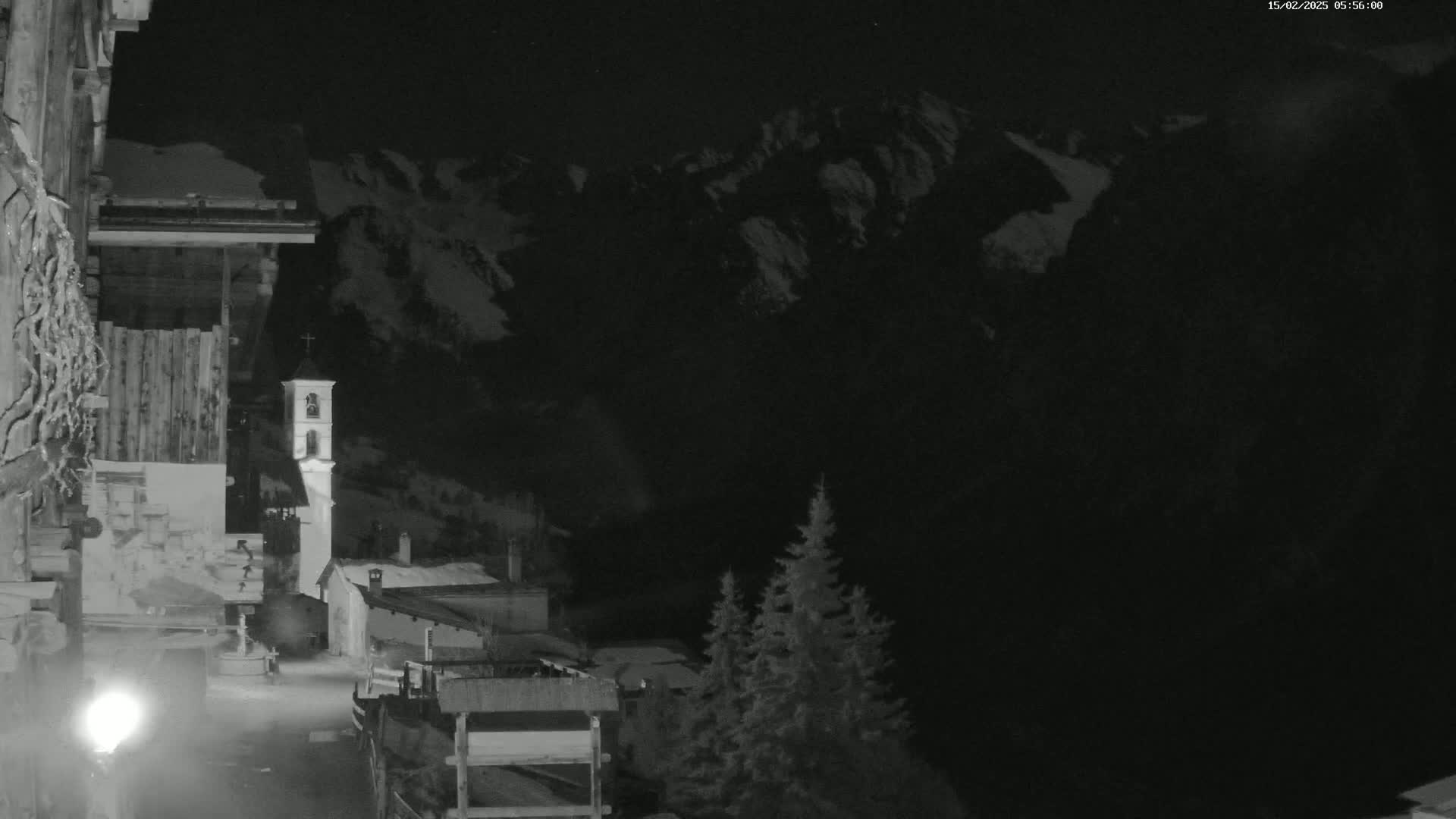A nighttime, snow-covered mountain range looms behind a small village with a tall, slender bell tower, illuminated by a nearby light source.