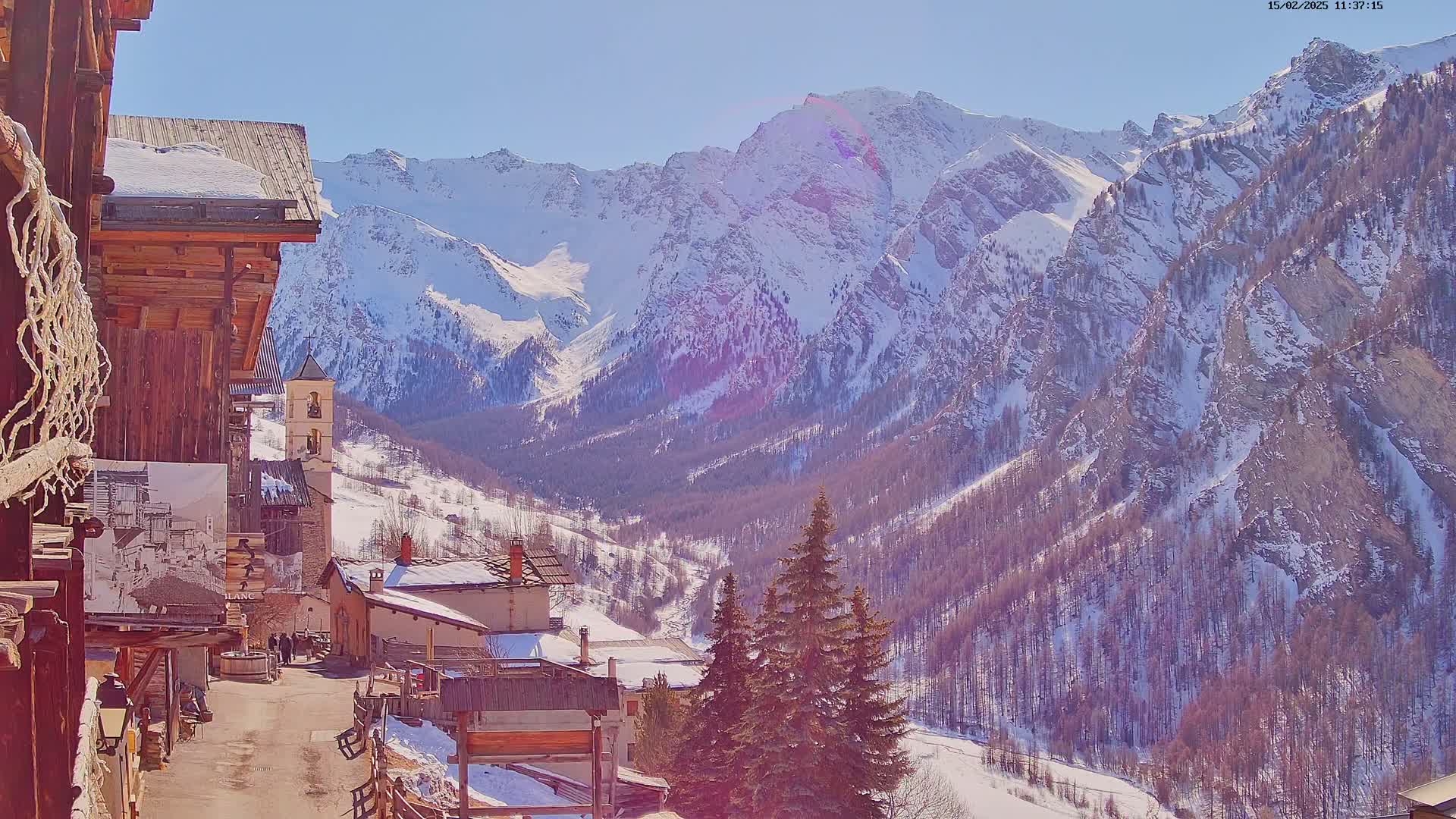 A snow-covered mountain village nestled in a valley, with a backdrop of towering, snow-capped mountains under a sunny sky.