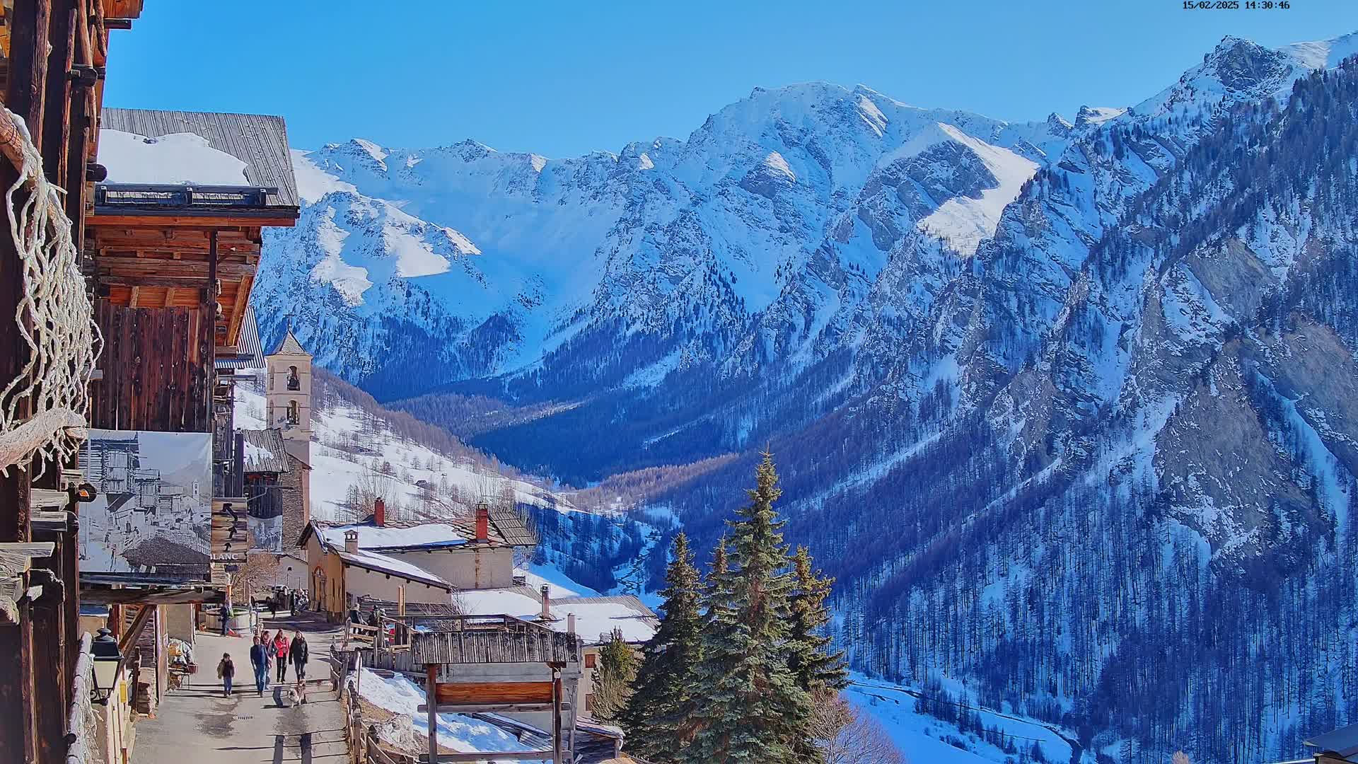A snow-covered mountain valley is viewed from a village of wooden buildings on a sunny day, with several people walking on a path.
