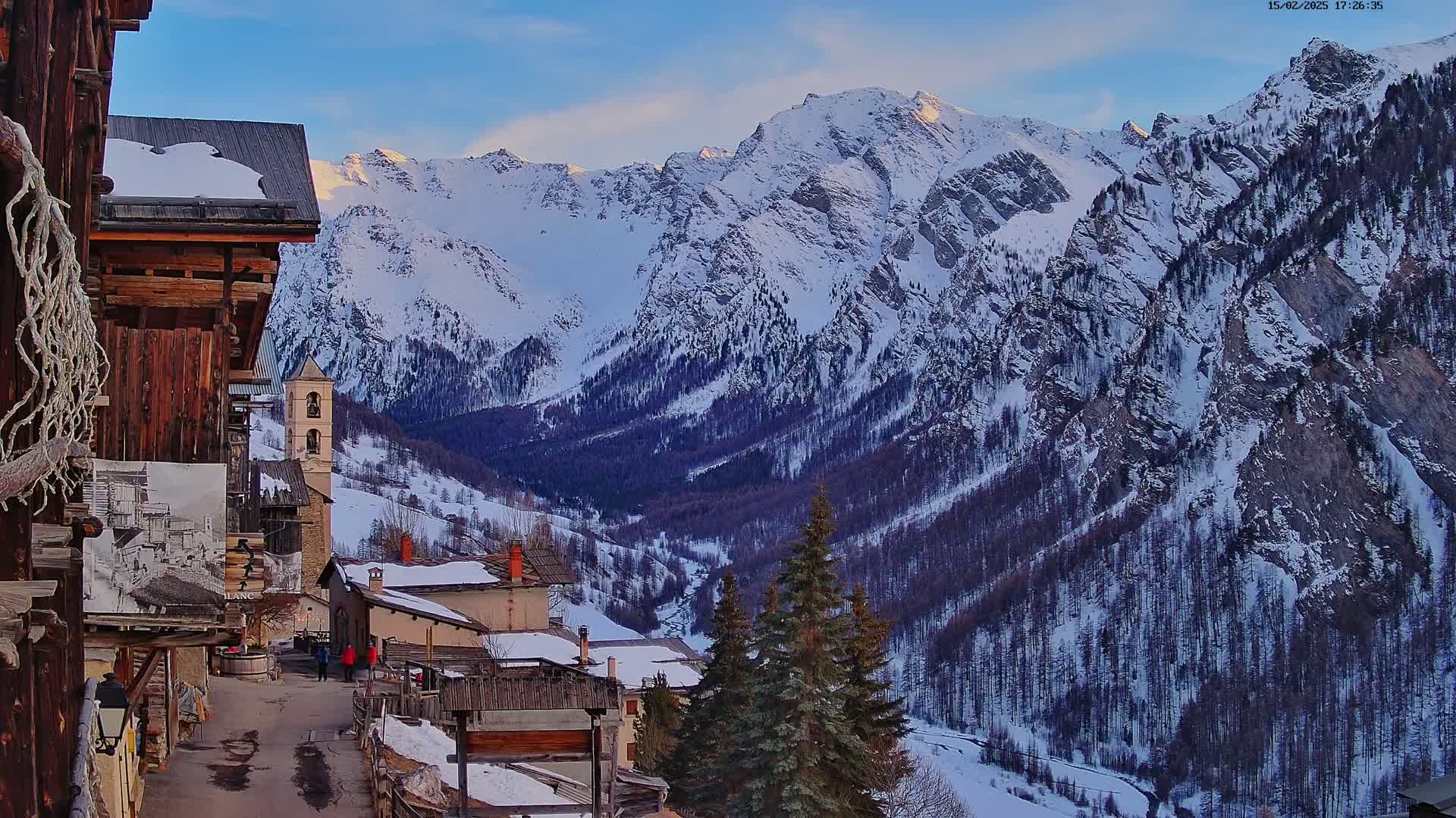 A snow-covered mountain valley at sunset, viewed from a village with old wooden buildings.