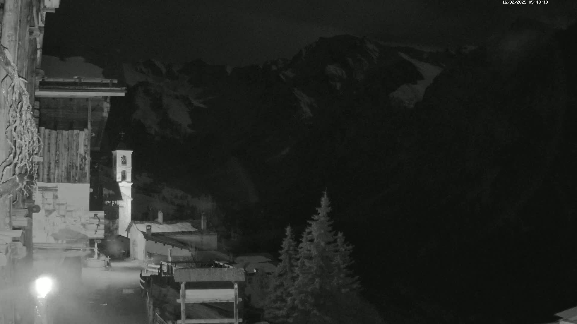 A nighttime, snow-covered mountain village scene shows a church tower illuminated, with evergreen trees in the foreground under a dark sky.