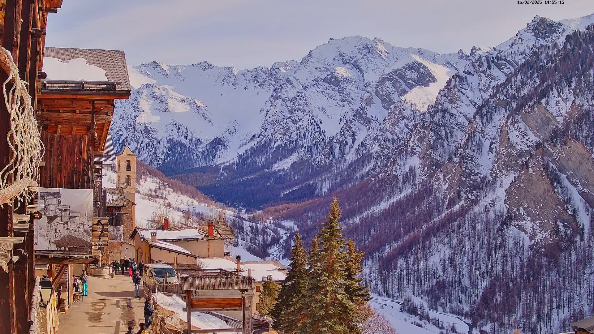A sunny day reveals a snow-covered mountain range overlooking a quaint village with traditional wooden buildings and a church.