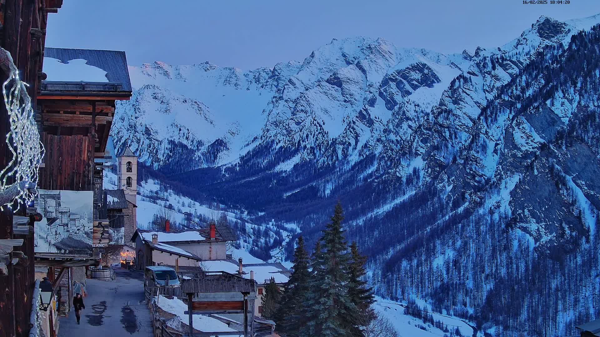 A snow-covered mountain village at dusk, nestled in a valley surrounded by towering snow-capped mountains.