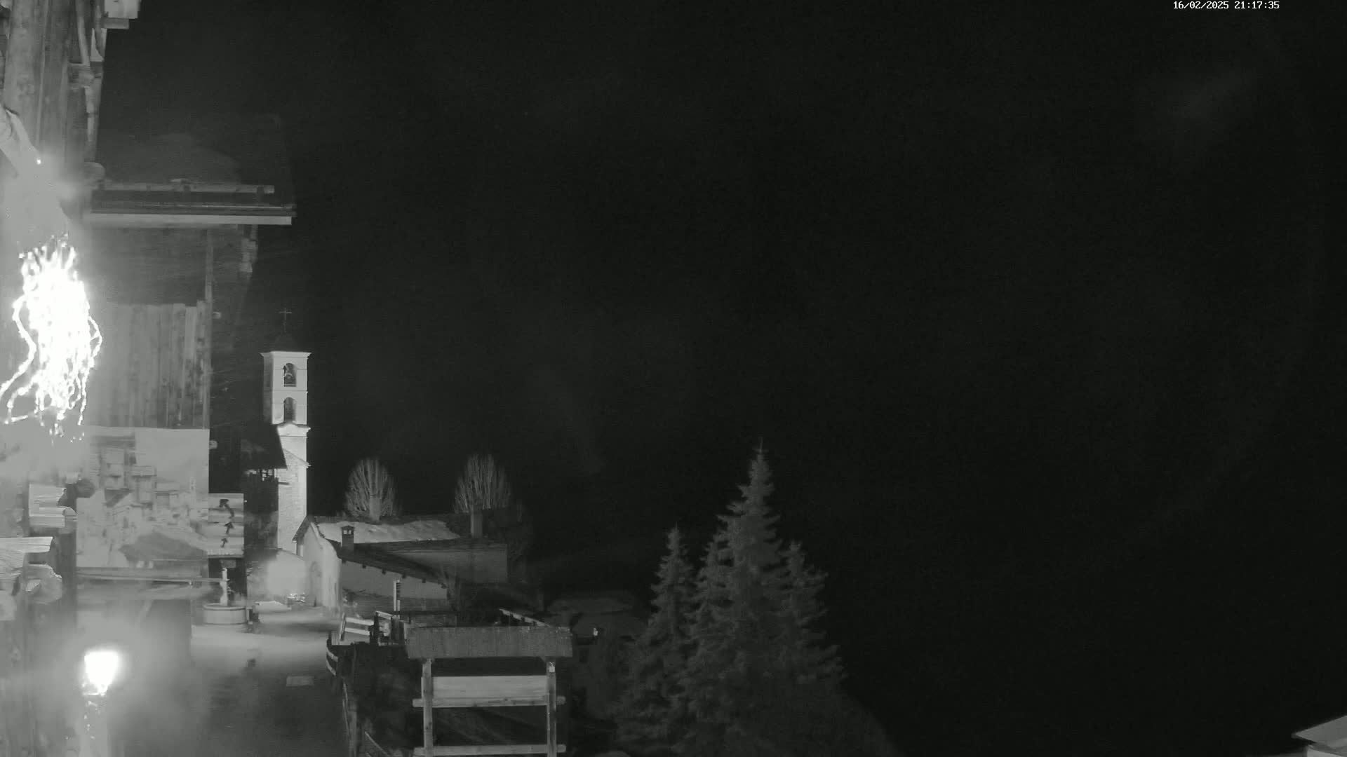 A nighttime, snowy scene shows a village with a bell tower, illuminated by fireworks, under a dark sky.