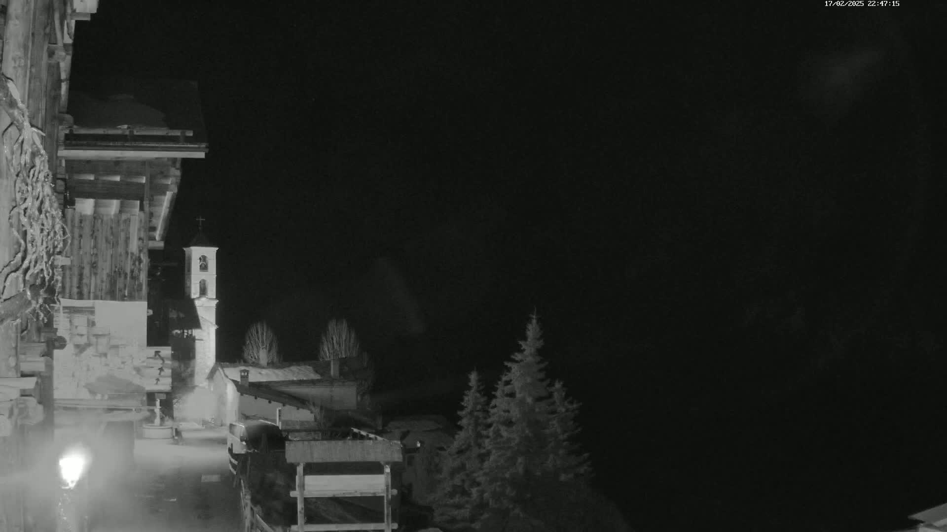 A nighttime, black-and-white view of a snow-covered village with a church steeple, partially obscured by darkness and a few evergreen trees.