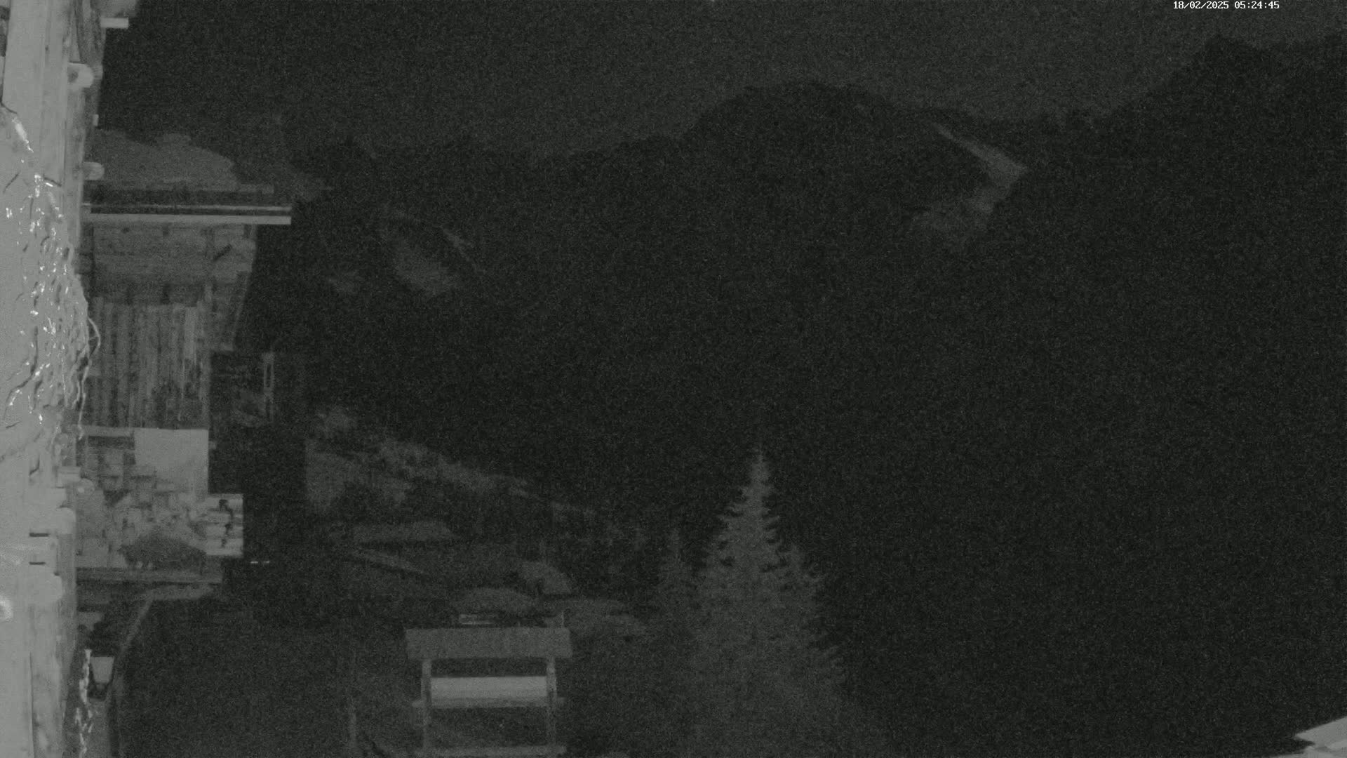 A nighttime, low-light image shows a wooden structure, possibly a building, with dark mountains in the background under a clear, dark sky.