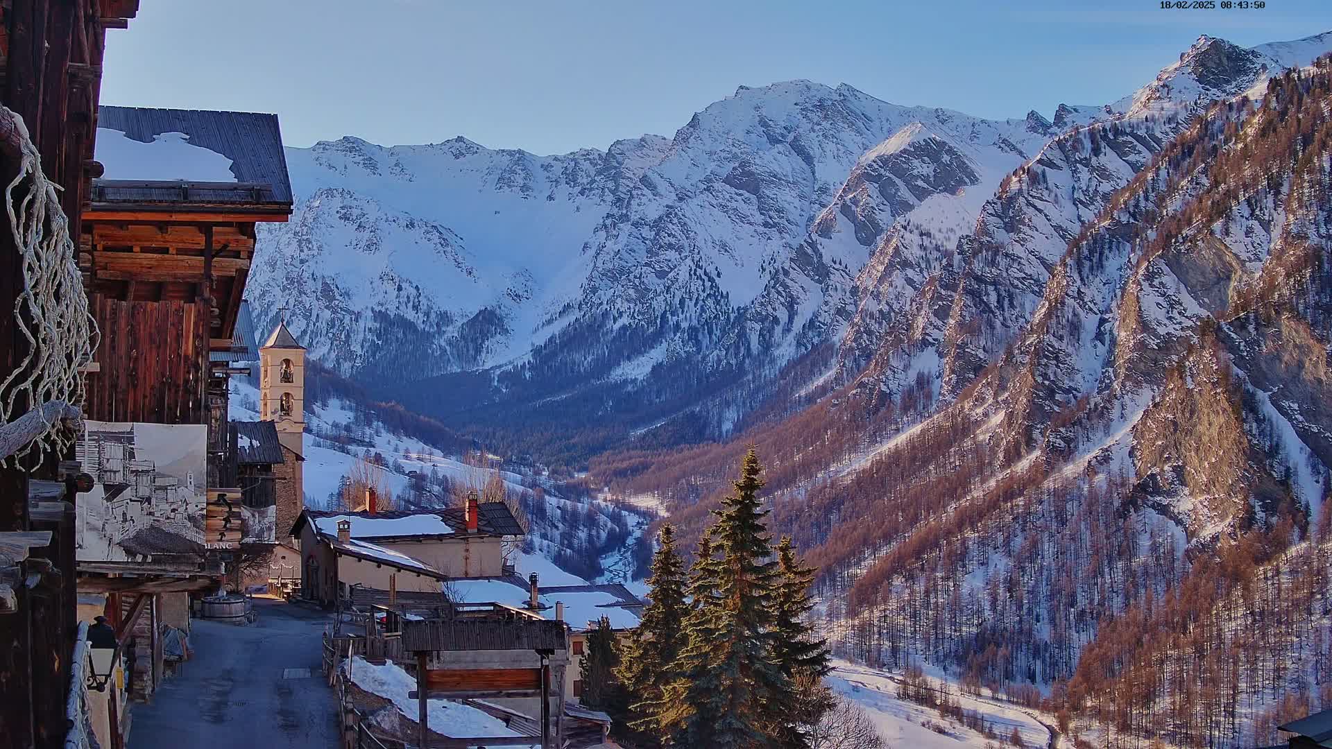 A snow-covered mountain valley is seen from a village of rustic buildings, with evergreens in the foreground under a clear sky.