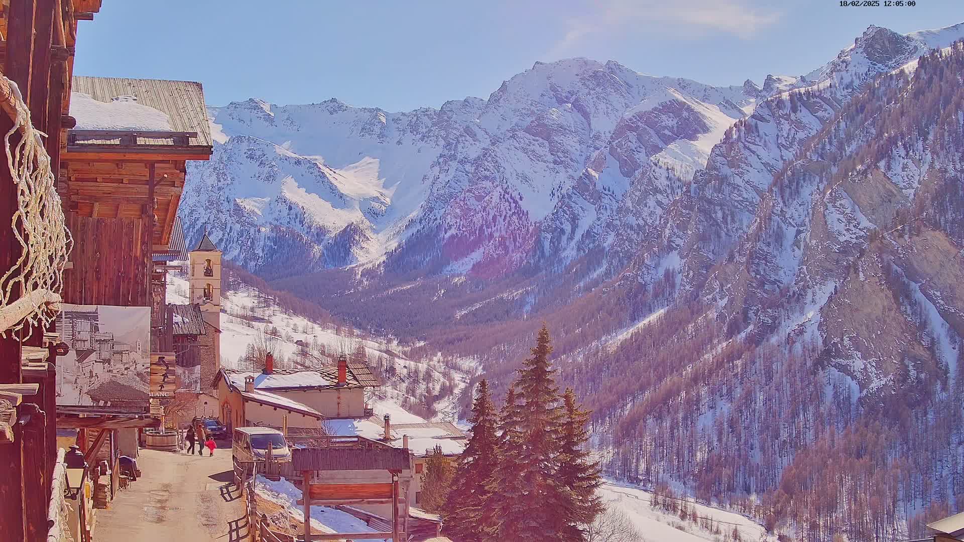 A sunny, snow-covered mountain valley scene with a small village nestled among the snow-dusted trees and towering, snow-capped mountains.