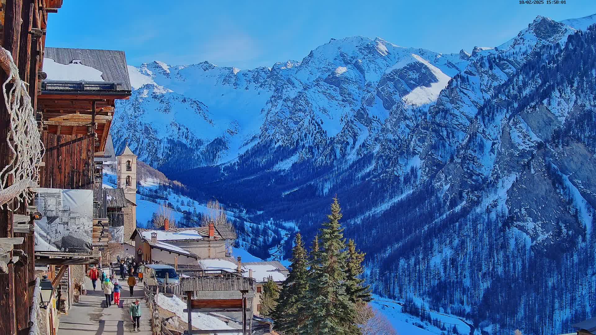 A snow-covered mountain range is visible behind a small village with wooden buildings and people walking on a pathway on a sunny day.