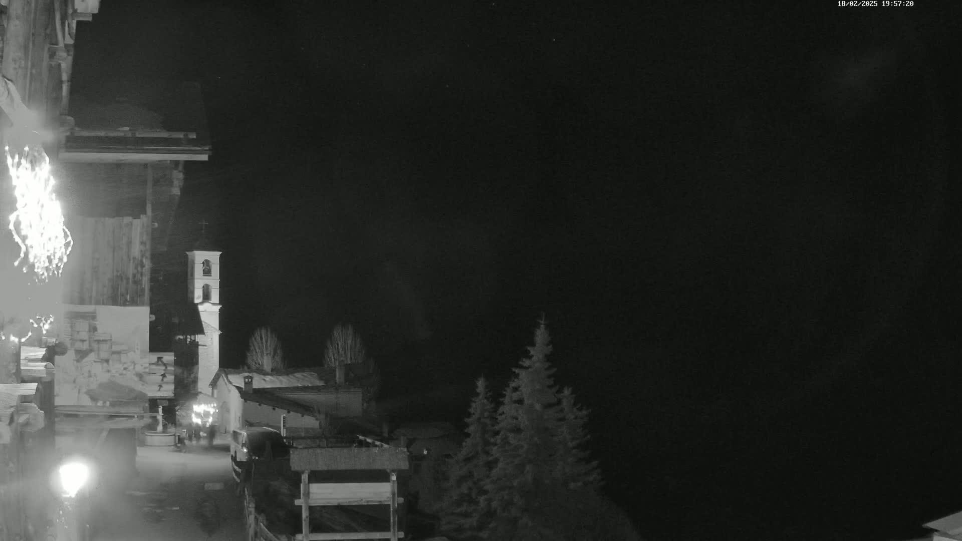 A nighttime view of a village with a bell tower, some vehicles, and evergreens under a dark sky.