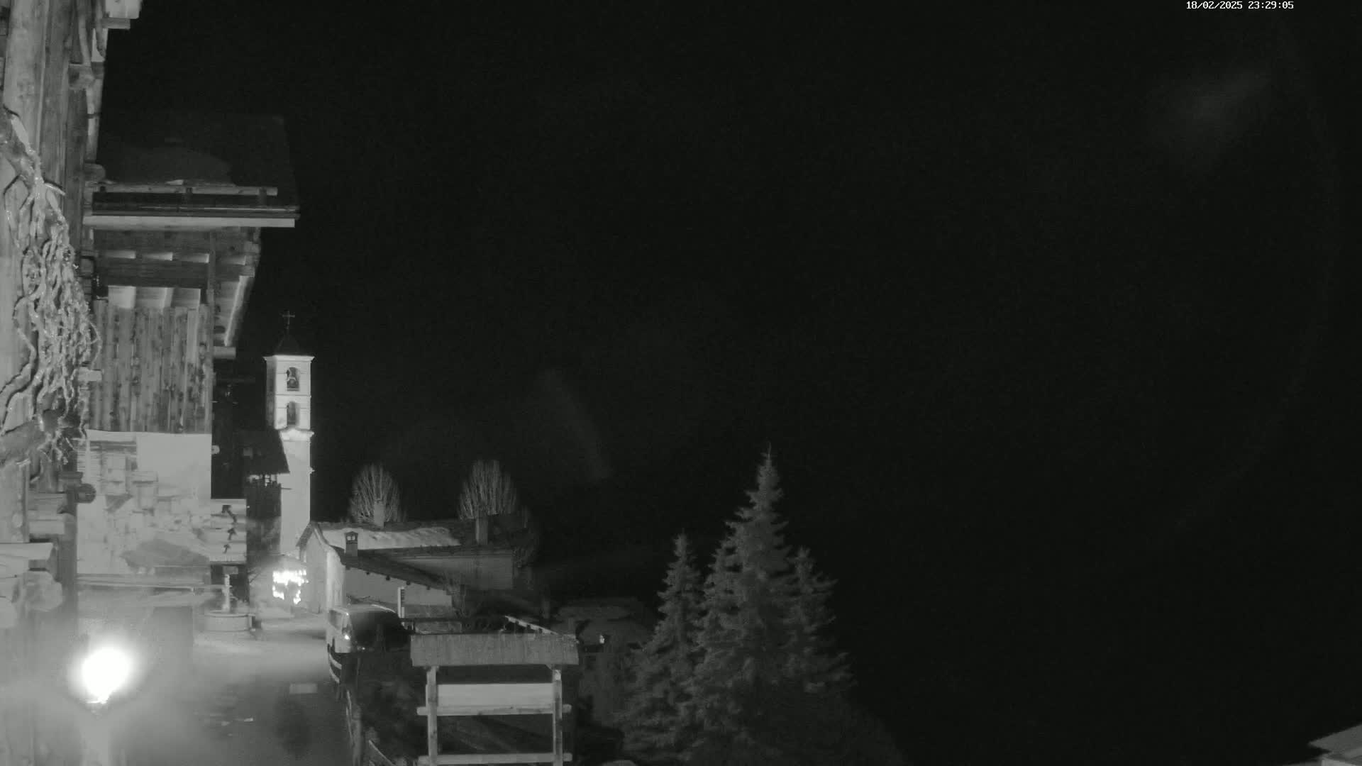 A nighttime, grayscale image shows a village scene with a tall, slender bell tower, some buildings, evergreen trees, and a dark, clear sky.