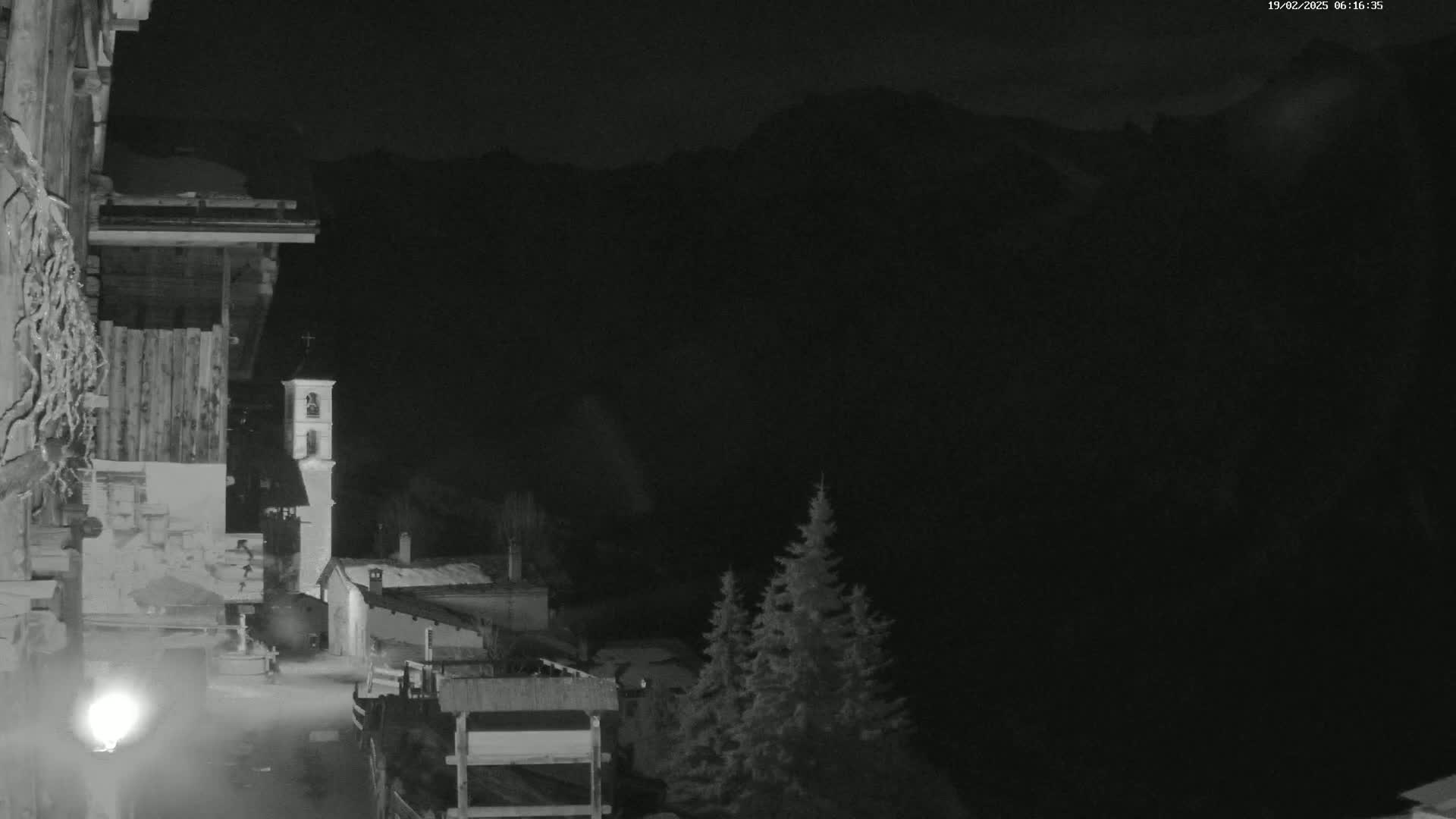 A nighttime, low-light view shows a small village nestled in a valley, with a church bell tower visible, and dark mountains looming in the background.